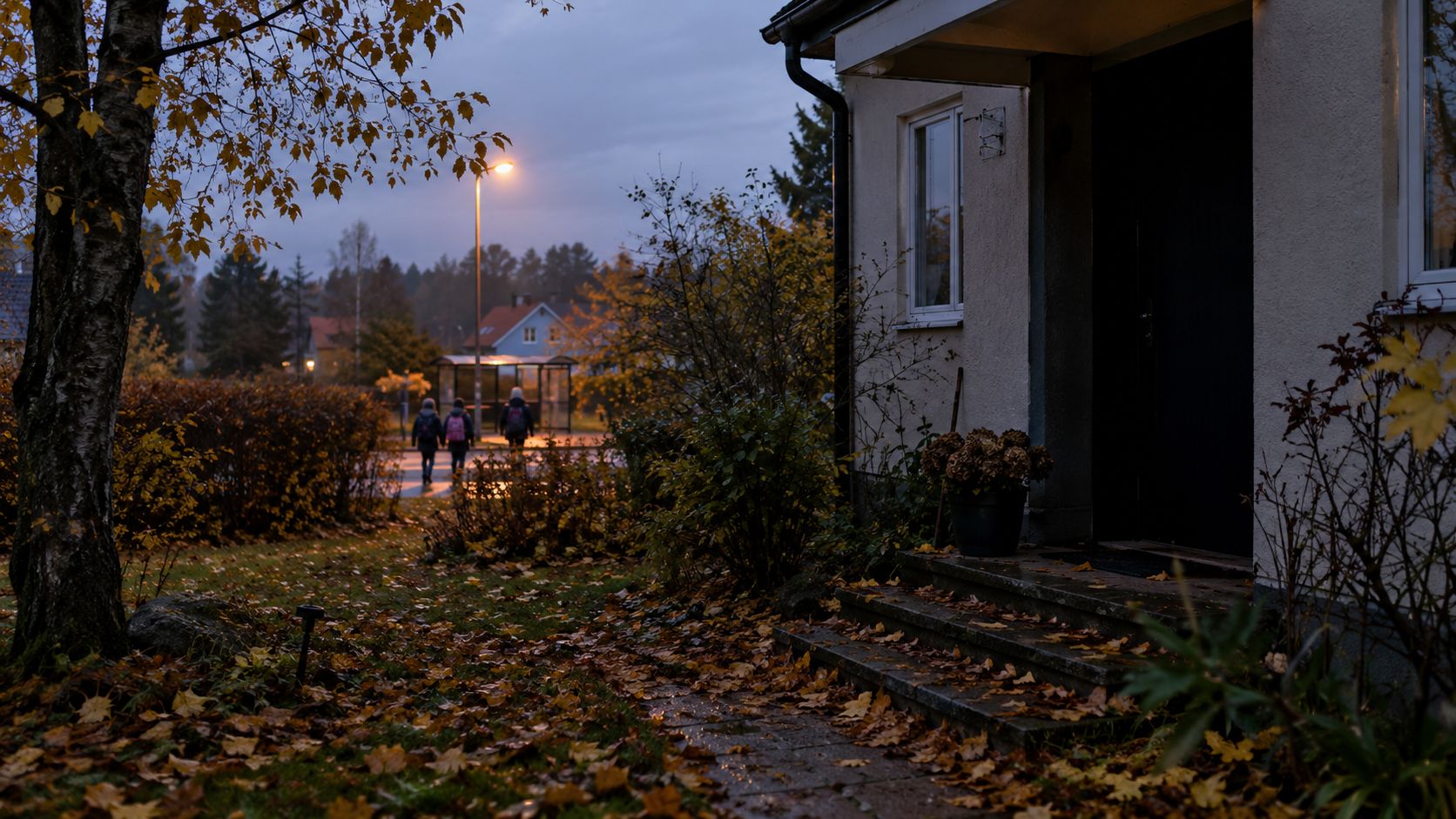 Höstig trädgård framför ett hus i skymningen med personer som går förbi på trottoaren i bakgrunden.